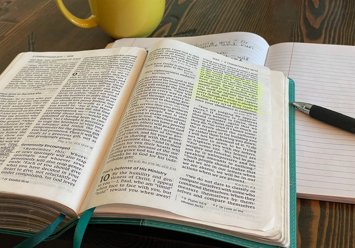 Bible laying open with teal colored book marks. Passage highlighted in yellow. Yellow coffee mug, notebook and pen in background